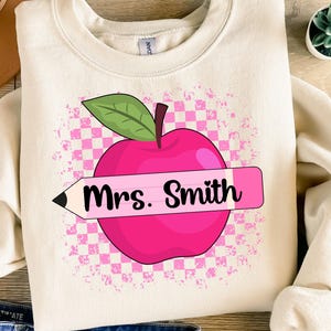 Peut inclure: Un sweat-shirt crème avec une pomme rose et un crayon rose avec le texte "Mme Smith" dessus. La pomme et le crayon sont sur un fond à carreaux rose et blanc.