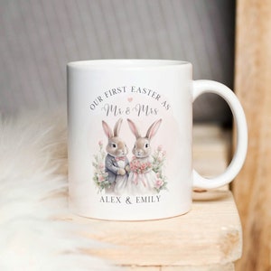 Op de afbeelding: Witte keramische mok met een roze en witte illustratie van twee konijnen die als bruid en bruidegom zijn gekleed. De tekst "Our First Easter as Mr & Mrs Alex & Emily" staat op de mok gedrukt.