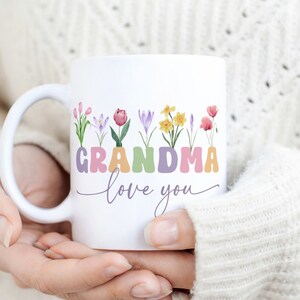 Puede incluir: Taza de cerámica blanca con un diseño floral colorido y el texto "Grandma love you".