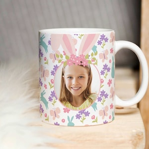 May include: White ceramic mug featuring a pink bunny ear design and a floral pattern. A girl's face is visible in a circular cutout on the mug.