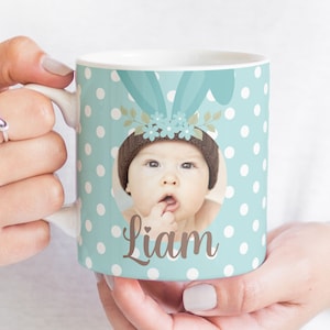 May include: A light blue ceramic mug with white polka dots. The mug features a photo of a baby wearing a brown hat with a floral headband and the name "Liam" printed below the photo.