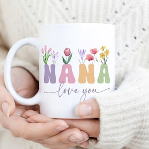 Puede incluir: Taza de cerámica blanca con un diseño floral que deletrea "NANA" en colores pastel. El texto "love you" está escrito en cursiva debajo del diseño floral.