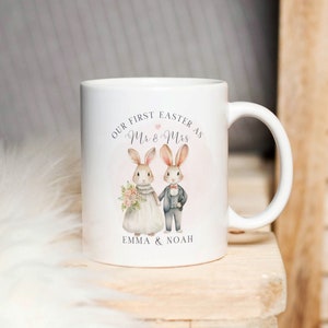 Op de afbeelding: Witte keramische mok met een roze en witte illustratie van twee konijnen die als bruid en bruidegom zijn gekleed. De tekst "Our First Easter as Mr & Mrs Emma & Noah" staat op de mok gedrukt.