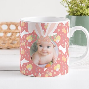May include: A white ceramic mug with a pink background and a pattern of white bunnies, yellow flowers, and colourful Easter eggs. A circle in the centre of the mug features a photo of a baby wearing a flower crown.