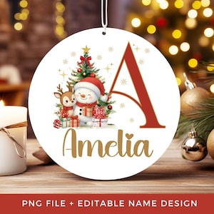 Pode incluir: Um enfeite de Natal redondo e branco com uma letra A vermelha grande, um boneco de neve, uma rena e presentes sob uma árvore decorada. O nome Amelia está impresso em letras douradas sob a ilustração e possui um cordão branco.