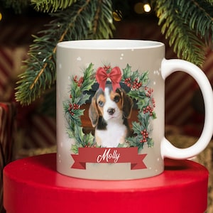Może przedstawiać: Kubek ceramiczny w kolorze beżowym z wzorem świątecznego wieńca z szczenięciem beagle, czerwoną kokardką i ostrokrzewem. Kubek ma biały uchwyt, a na czerwonej wstążce nadrukowano imię "Molly".