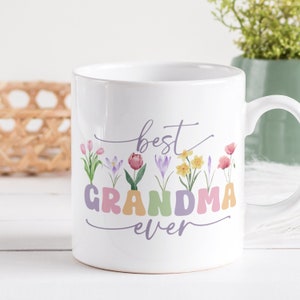May include: White ceramic mug with the words "best GRANDMA ever" in pastel colors. The letters are decorated with colorful flowers. The mug has a white handle and is on a white surface.
