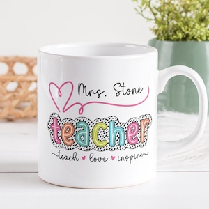 Puede incluir: Taza de cerámica blanca con un corazón rosa y el texto "Mrs. Stone teacher teach love inspire" en letras de colores con lunares negros.