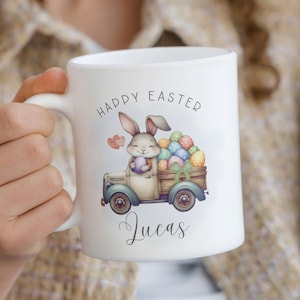 Può includere: Tazza in ceramica bianca con un coniglio dei cartoni animati che trasporta un cesto di uova di Pasqua in un camion d'epoca. Il testo "Happy Easter" è sopra il coniglio e il nome "Lucas" è sotto il camion.