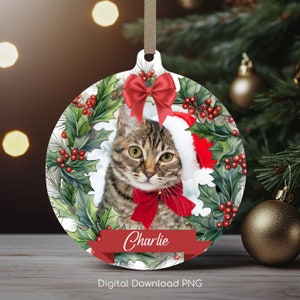 May include: A round Christmas ornament featuring a cat wearing a Santa hat and a red bow. The cat is surrounded by a green holly wreath with red berries. The ornament has the name "Charlie" written on a red ribbon.