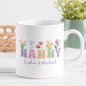 Puede incluir: Taza de cerámica blanca con un diseño floral que deletrea "NANNY" en letras de colores. La taza tiene los nombres "Sophia & Michael" escritos debajo del diseño floral.