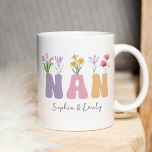 Puede incluir: Taza de cerámica blanca con un diseño floral y la palabra "NAN" en colores pastel. La taza también tiene el texto "Sophia & Emily" impreso en la parte inferior.