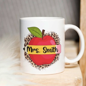 May include: White ceramic mug with a red apple and a yellow pencil with the text "Mrs. Smith" on it. The apple is surrounded by a brown and black leopard print.