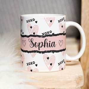 May include: White ceramic mug with a pink and black heart pattern. The mug has a black band with the name "Sophia" in cursive script and a pink background.