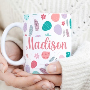 May include: White ceramic mug with a pink, green, and purple Easter egg pattern. The mug has the name "Madison" written in pink cursive.