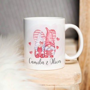 以下が含まれることがあります： 赤と白のストライプのハートのデザインが施された白いセラミックマグカップ。2人のノームがハートを持っています。1つは赤、もう1つは白です。マグカップには「Camila & Oliver」という文字が印刷されています。