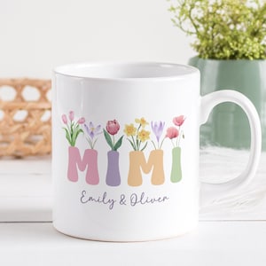 Puede incluir: Taza de cerámica blanca con la palabra "MIMI" escrita en letras de colores con flores en cada letra. El texto "Emily & Oliver" está impreso debajo de las letras.