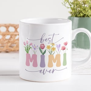 Puede incluir: Taza de cerámica blanca con un diseño floral y el texto "best mimi ever". Las flores son de diferentes colores, incluyendo rosa, morado, amarillo y verde.