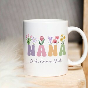 Puede incluir: Taza de cerámica blanca con un diseño floral que deletrea "NANA" en letras de colores. La taza tiene un mensaje personalizado en la parte inferior que dice "Zack, Emma, Noah".