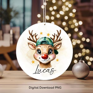 May include: A round Christmas ornament featuring a cartoon reindeer wearing a green baseball cap. The reindeer has a red nose and big brown eyes. The ornament is white with gold stars and the name "Lucas" written in black cursive.