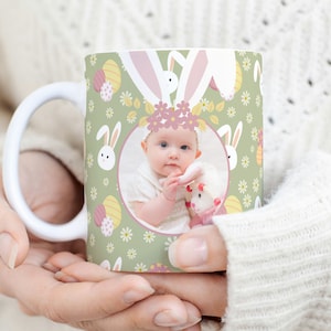 May include: A white ceramic mug with a green background and a pattern of Easter eggs, bunnies, and flowers. A photo of a baby girl wearing a pink headband is in the centre of the mug.