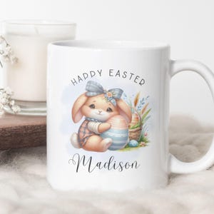 May include: White ceramic mug with a colorful illustration of a bunny holding an Easter egg. The mug says "Happy Easter" and "Madison" in a cursive font.