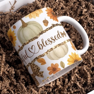 May include: White ceramic mug with a fall-themed design. The mug features pumpkins, sunflowers, and the words "grateful blessed" in a gold script. The design includes a gold glitter band. The mug is in a box with brown paper.