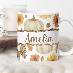 Op de afbeelding: Een witte keramische mok met een herfstpatroon van lichtgroene pompoenen, gele zonnebloemen, bruine eikels en een strik met luipaardprint. Een centrale horizontale band met gouden glitterranden toont de naam Amelia in bruin schrift.