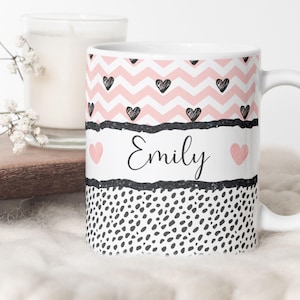 Op de afbeelding: Witte keramische mok met een roze en zwart chevronpatroon, zwarte harten en een zwart-wit stippenpatroon. De mok heeft de naam "Emily" in zwarte, cursieve letters.