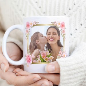 May include: White ceramic mug with a gold frame and floral design. Inside the frame is a photo of a mother and daughter. The daughter is kissing the mother on the cheek. The photo is surrounded by pink flowers.