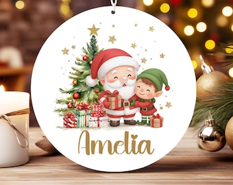 May include: A round white Christmas ornament featuring a watercolor illustration of Santa Claus and an elf holding a gift in front of a decorated pine tree with gold stars. The name Amelia is printed in gold script at the bottom of the ornament.