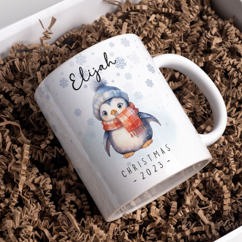 May include: White ceramic mug featuring a penguin wearing a blue hat and red scarf. The mug is personalised with the name "Elijah" and the text "Christmas - 2023".