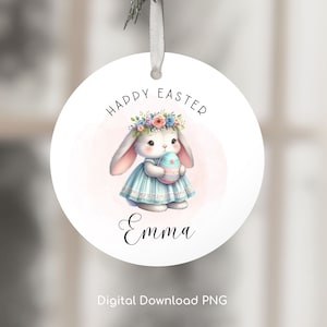 May include: A white round ornament with a pink background and a cartoon bunny holding a decorated Easter egg. The bunny is wearing a blue and white dress and a flower crown. The text "Happy Easter" is above the bunny and the name "Emma" is below the bunny.