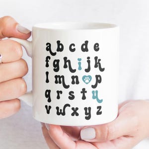 May include: White ceramic mug with a black alphabet printed on it. The letter "i" is replaced with a blue paw print inside a heart.