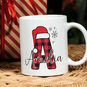 以下が含まれることがあります： 白のセラミックマグ。赤と黒のバッファローチェックの「A」の文字にサンタ帽のデザイン。文字の下には筆記体で「Amelia」と書かれています。マグの右側には雪の結晶のグラフィックがあります。背景にはギフトボックスがあります。