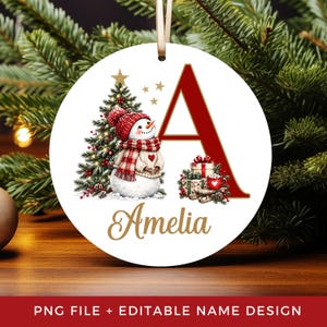May include: A round white Christmas ornament featuring a large red letter A, a snowman wearing a red knit hat and scarf, a decorated Christmas tree, wrapped gifts, and the name Amelia in gold script. A beige ribbon is attached to the top.