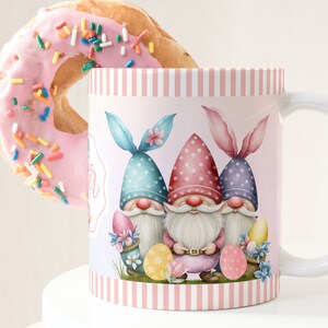May include: White ceramic mug with a pink and white striped design. The mug features a colorful illustration of three gnomes holding Easter eggs. The text "Happy Easter" is visible on the mug.