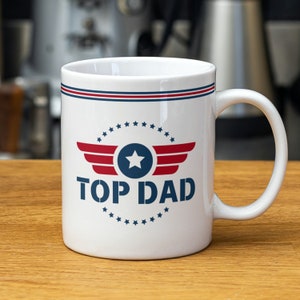 Puede incluir: Taza de café de cerámica blanca con un diseño rojo, blanco y azul. La taza presenta una estrella y alas con el texto "TOP DAD" en una fuente de esténcil azul y blanca.
