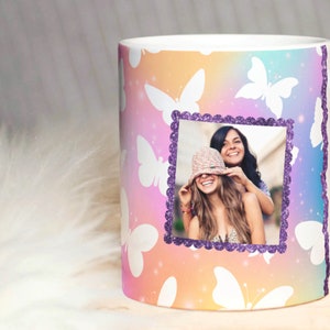 Puede incluir: Taza de cerámica blanca con un fondo degradado de colores y un patrón de mariposa blanca. Una foto de dos mujeres está en un marco de purpurina morado.