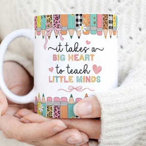 Op de afbeelding: Witte keramische mok met de tekst "it takes a BIG HEART to teach LITTLE MINDS". De mok heeft een kleurrijk potloodontwerp rond de rand, met roze strikken en harten. Een cadeau voor leraren.