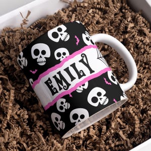 Può includere: Tazza in ceramica bianca con un motivo a teschi bianco e nero e uno striscione rosa con il nome "EMILY" stampato.