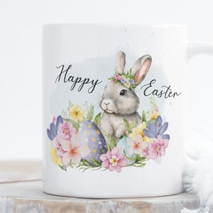 Puede incluir: Taza de cerámica blanca con un conejo gris que lleva una corona de flores y está rodeado de flores coloridas y huevos de Pascua. El texto "Happy Easter" está impreso en la taza.