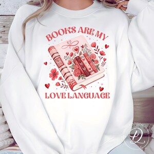 May include: White sweatshirt with the text "BOOKS ARE MY LOVE LANGUAGE" in red. The graphic features a pink book with floral and heart accents. Additional red hearts and flowers are scattered around the design.