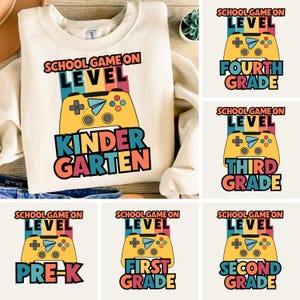 Puede incluir: Sudadera crema con un gráfico colorido que presenta un mando de videojuegos amarillo y las palabras "SCHOOL GAME ON LEVEL KINDERGARTEN". Imágenes adicionales muestran diseños similares para otros grados.