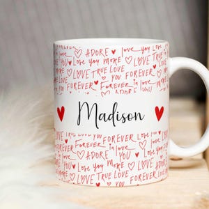 Op de afbeelding: Een witte keramische mok met een rood en zwart ontwerp. De mok heeft de woorden "love", "adore", "true love" en "forever" die in een handschrift-stijl worden herhaald. De naam "Madison" is in zwarte letters geschreven met een rood hart aan beide kanten.