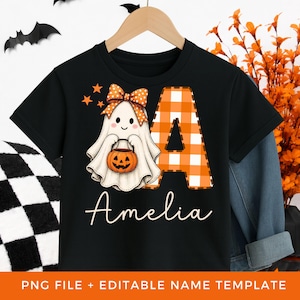 Puede incluir: Una camiseta negra de manga corta con un gráfico de un fantasma blanco con un lazo naranja de lunares, sosteniendo un cubo de calabaza naranja. Junto al fantasma hay una letra A grande de cuadros naranjas y blancos, con el nombre Amelia debajo.