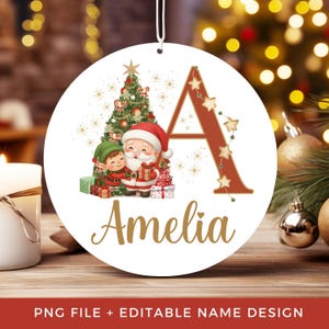 Pode incluir: Um enfeite de Natal redondo e branco com uma letra A vermelha grande decorada com estrelas, um Papai Noel e um elfo de desenho animado ao lado de uma árvore de Natal com presentes, e o nome Amelia escrito em letras douradas abaixo.