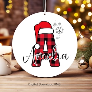 Peut inclure: Une boule ronde blanche avec une lettre A en tissu écossais rouge et noir surmontée d'un chapeau de Père Noël rouge. Le nom Amelia est écrit en cursive noire sous la lettre. La boule est décorée de flocons de neige.
