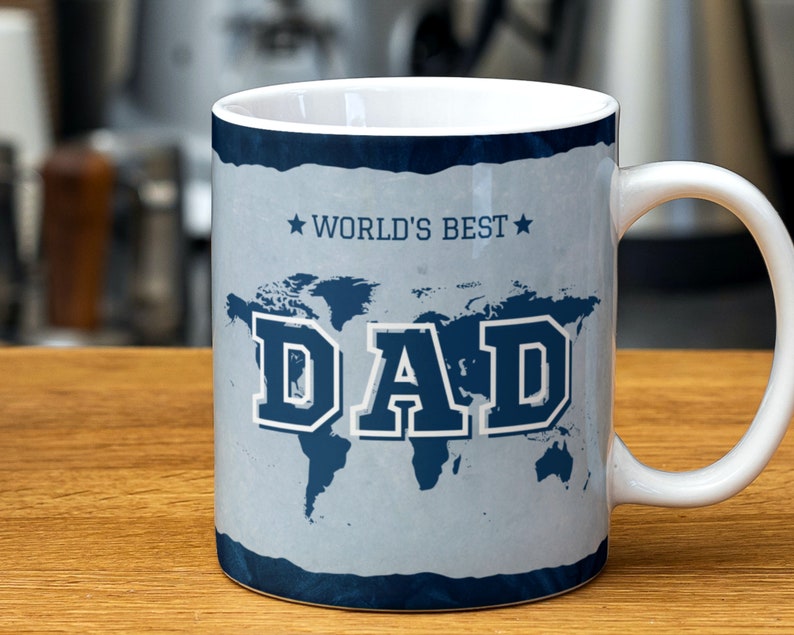May include: White ceramic mug with a blue and gray design. The mug features a world map with the word "DAD" in large, bold, blue letters. The text "WORLD'S BEST" is printed in smaller blue letters above the map.