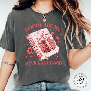 May include: Dark grey t-shirt with the text "BOOKS ARE MY LOVE LANGUAGE" in pink. The graphic features a pink book with floral and heart accents. The design includes a stack of books and decorative flowers.